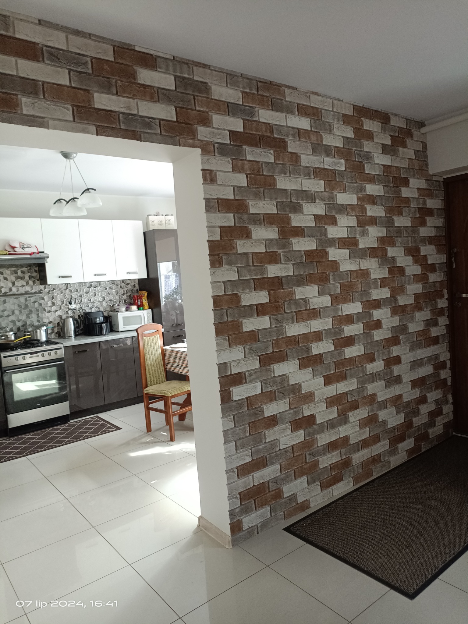 Ściana w kuchni wykończona dekoracyjną okładziną imitującą cegłę w odcieniach szarości, brązu i bieli, widok na wnętrze kuchni z białymi szafkami i sprzętami AGD, jasna podłoga z płytek.