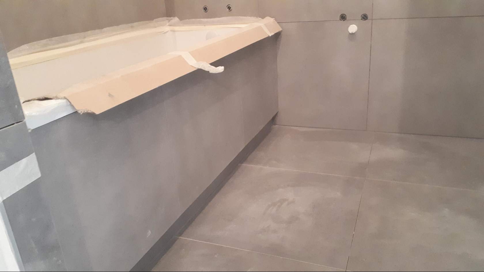 Szara łazienka w trakcie wykańczania: obudowana wanna z zabezpieczeniami kartonowymi, szare płytki na ścianach i podłodze, widoczne punkty instalacji.