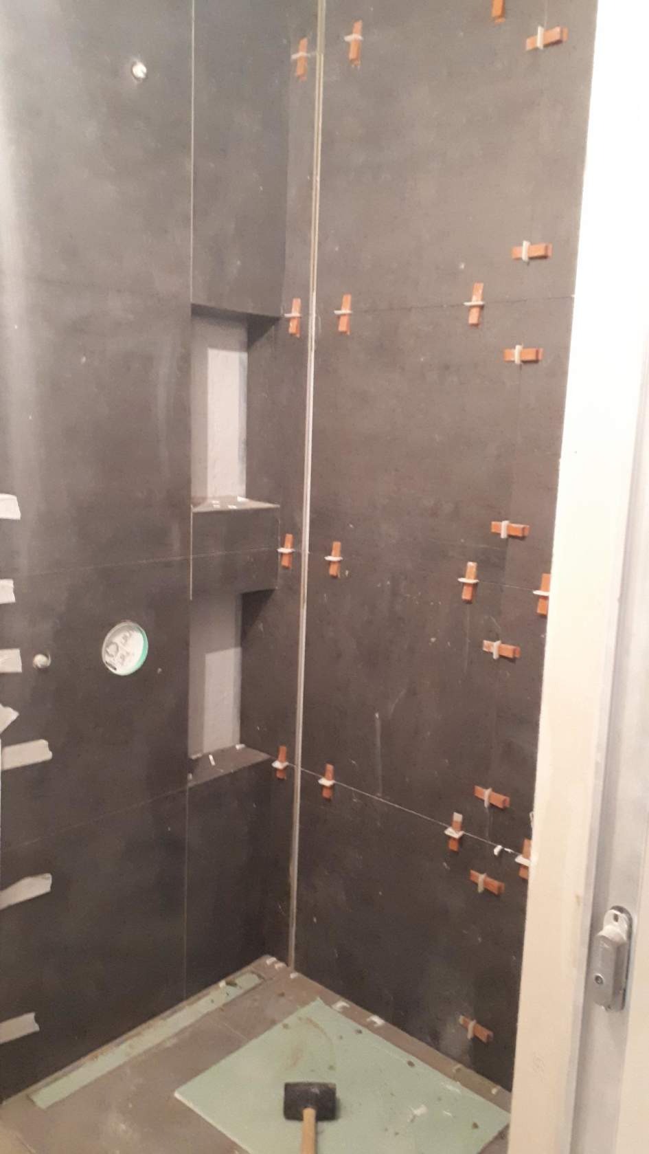 Ciemne płytki ścienne w trakcie układania w łazience, krzyżyki dystansowe, poziomica laserowa, przygotowane wnęki i podłoże, młotek gumowy na podłodze.