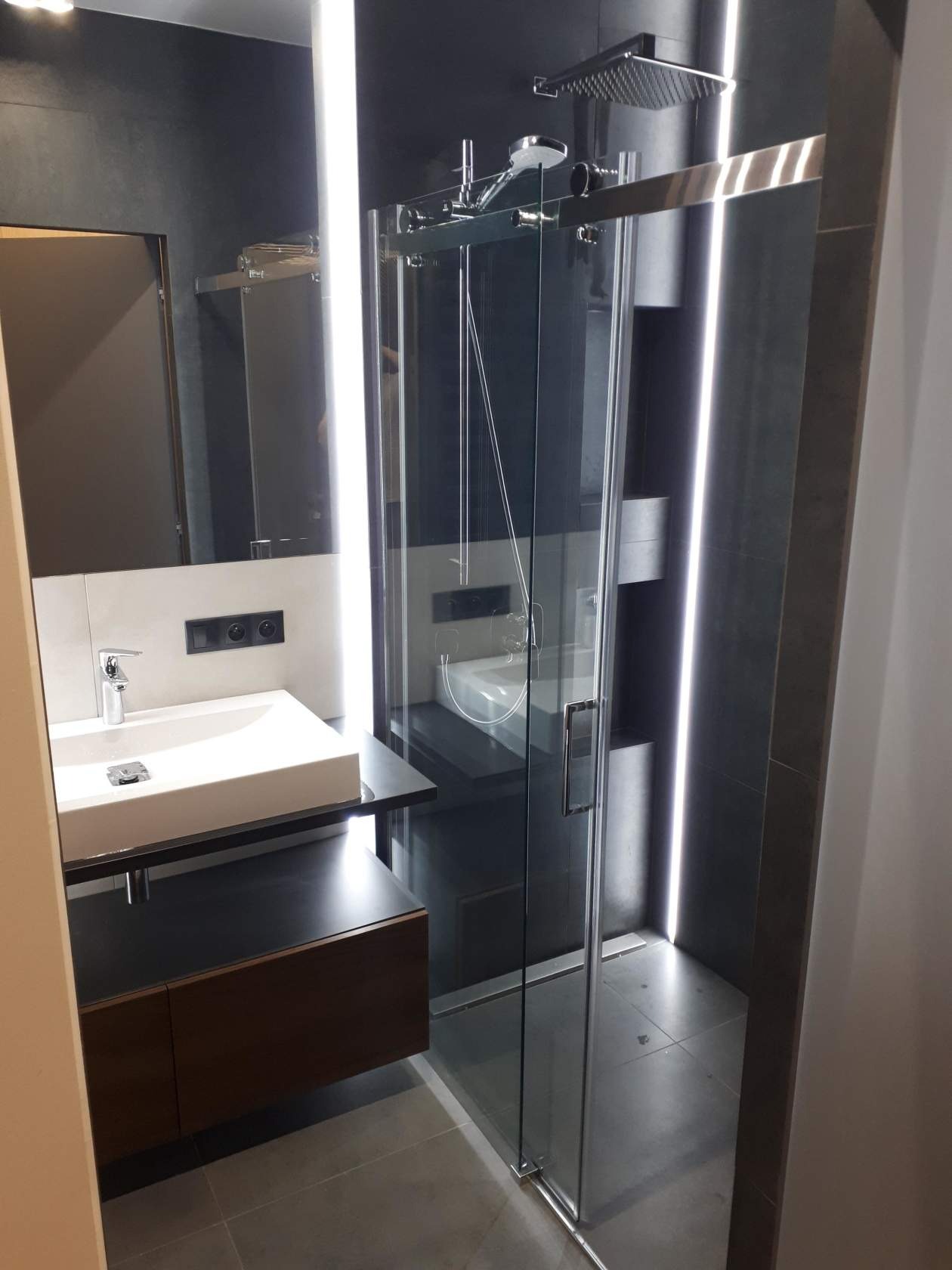 Nowoczesna łazienka z kabiną prysznicową ze szklanymi drzwiami przesuwnymi, białą umywalką nablatową na czarnym blacie i podświetleniem LED wzdłuż ściany w ciemnych odcieniach szarości.