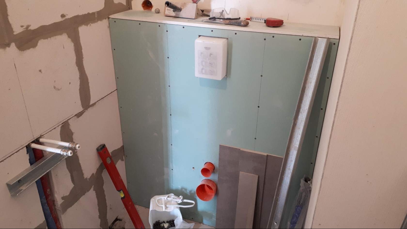 Stelaż zabudowy z płyt karton-gips w trakcie montażu, widoczne rury instalacji wodnej, poziomica, puszka instalacyjna Grohe, elementy kanalizacyjne i narzędzia.