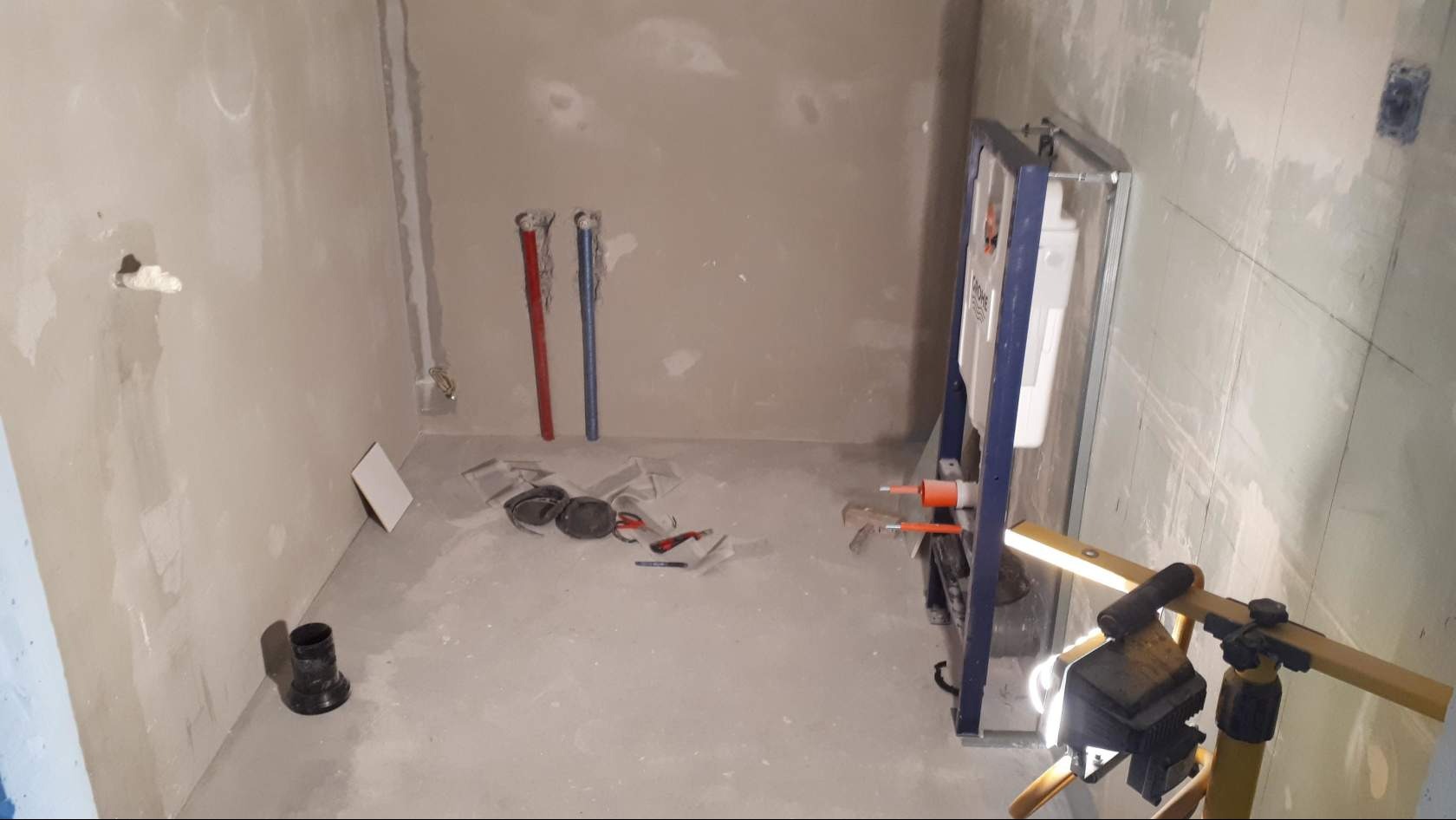 Surowa łazienka w trakcie prac: widoczne rury instalacyjne (czerwona i niebieska), stelaż podtynkowy WC Grohe, lampa robocza na statywie, narzędzia glazurnicze i fragment płytki na podłodze, ściany...