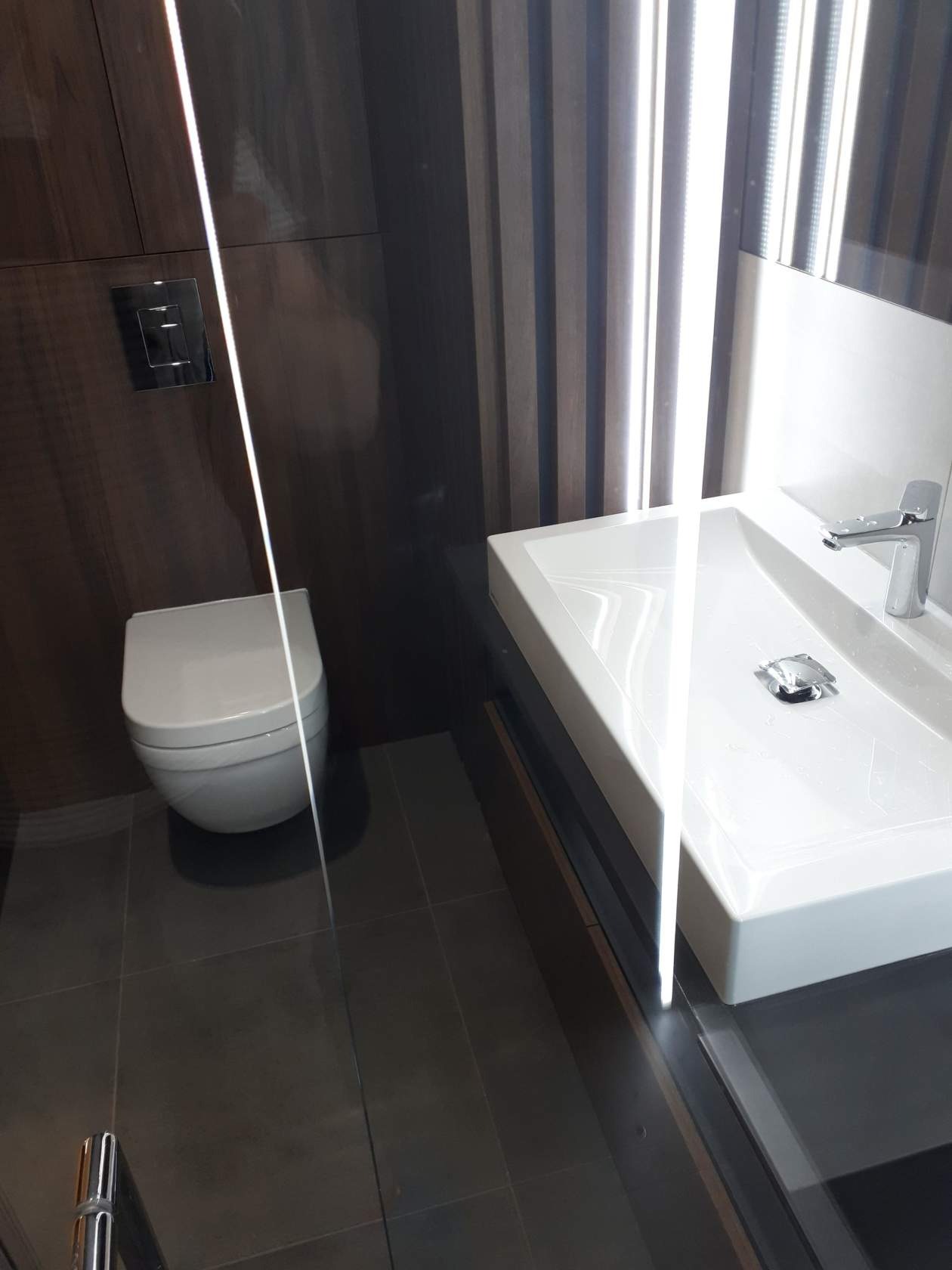 Nowoczesna łazienka z wiszącą toaletą, prostokątną umywalką nablatową z chromowaną armaturą i ciemnymi, drewnopodobnymi panelami ściennymi z oświetleniem LED.