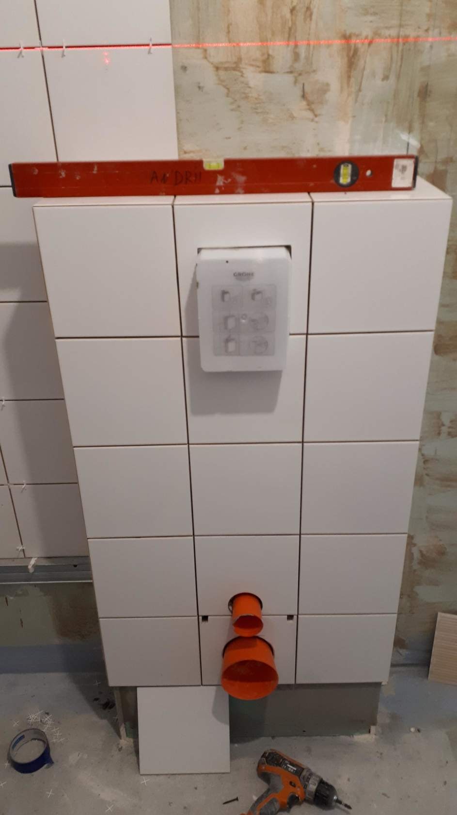 Białe płytki ceramiczne ułożone na ścianie z zamontowanym panelem sterującym i pomarańczową rurą odpływową, poziomica spoczywa na górnej krawędzi, na podłodze leży wkrętarka.