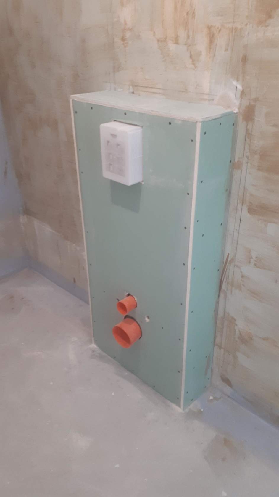 Stelaż podtynkowy obudowany płytą kartonowo-gipsową z widocznymi przyłączami kanalizacyjnymi i zamontowanym przyciskiem spłukującym, surowa ściana w tle.