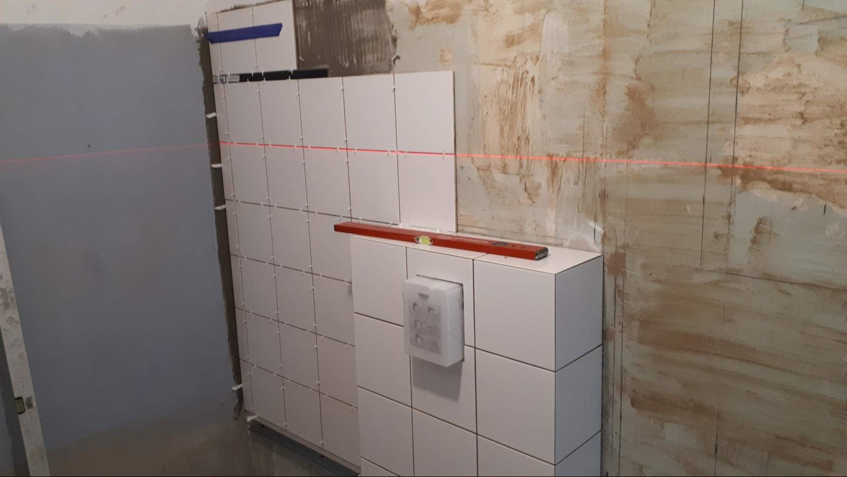 Układanie białych płytek w łazience, widoczny laserowy poziom, poziomica na zabudowie stelaża podtynkowego WC, ściana przygotowana pod płytki.