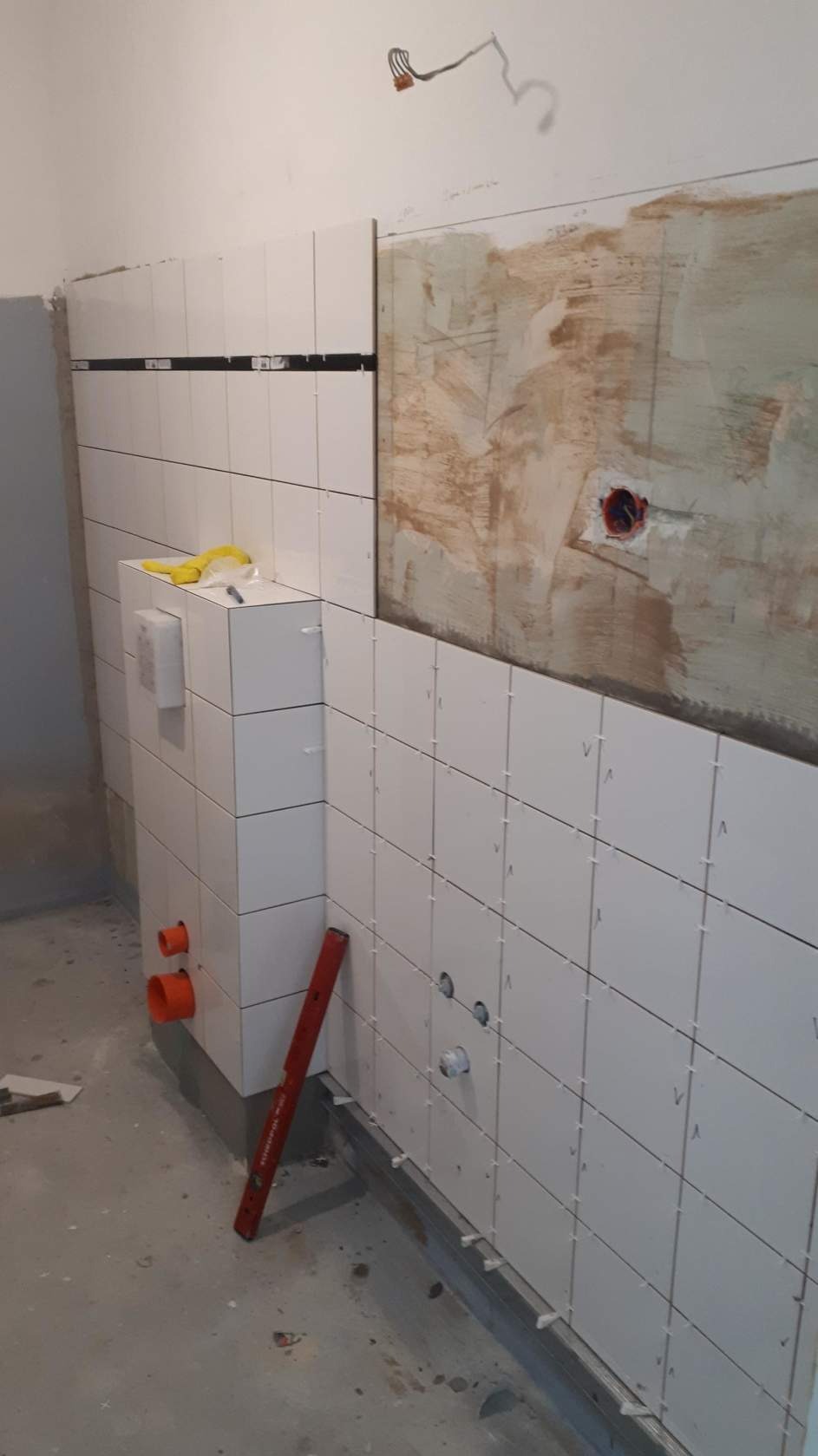 Ściana łazienki w trakcie układania białych płytek, widoczne rury instalacyjne, poziomica oparta o ścianę oraz fragment ściany przygotowanej pod płytki z widocznym otworem na gniazdko elektryczne.
