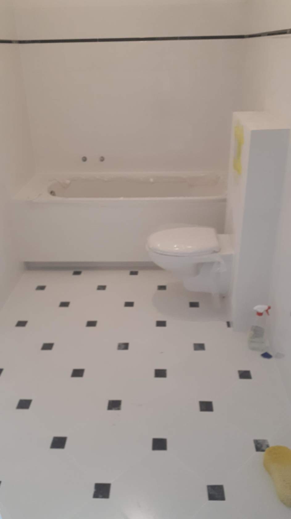 Biała łazienka po remoncie z nową toaletą, wanną zabezpieczoną folią i podłogą w biało-czarną szachownicę.