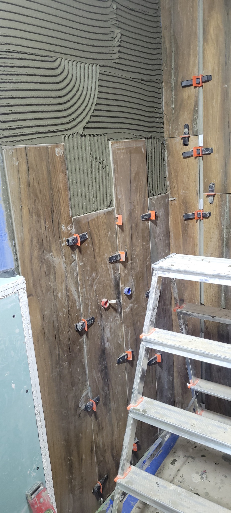 Układanie płytek imitujących drewno na ścianie z użyciem klipsów poziomujących; widoczny fragment ściany z nałożonym klejem w charakterystyczne paski oraz drabina aluminiowa.