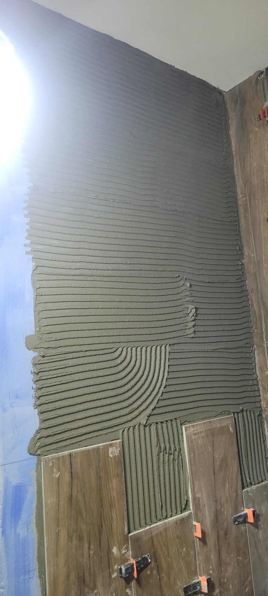 Ściana łazienki w trakcie układania płytek imitujących drewno, widoczne poziome pasy kleju i system poziomowania płytek.