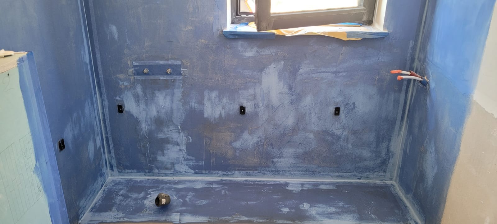 Niebieska hydroizolacja w trakcie wykonywania w łazience, widoczne przygotowane punkty elektryczne i instalacyjne, okno zabezpieczone folią.