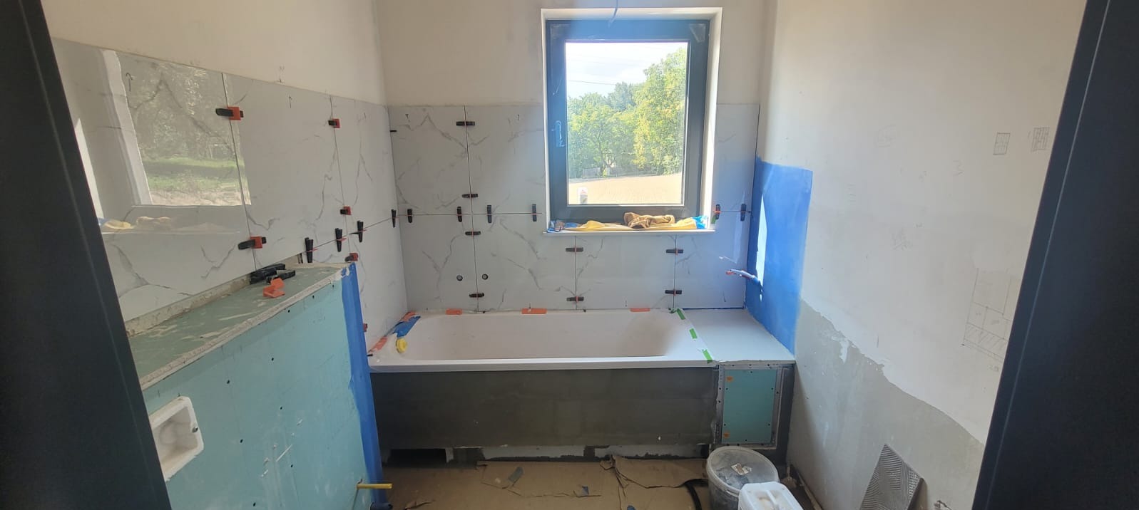 Wnętrze łazienki w trakcie układania płytek imitujących marmur, z widocznymi krzyżykami dystansowymi, poziomica na wannie i rysunek planu na ścianie.