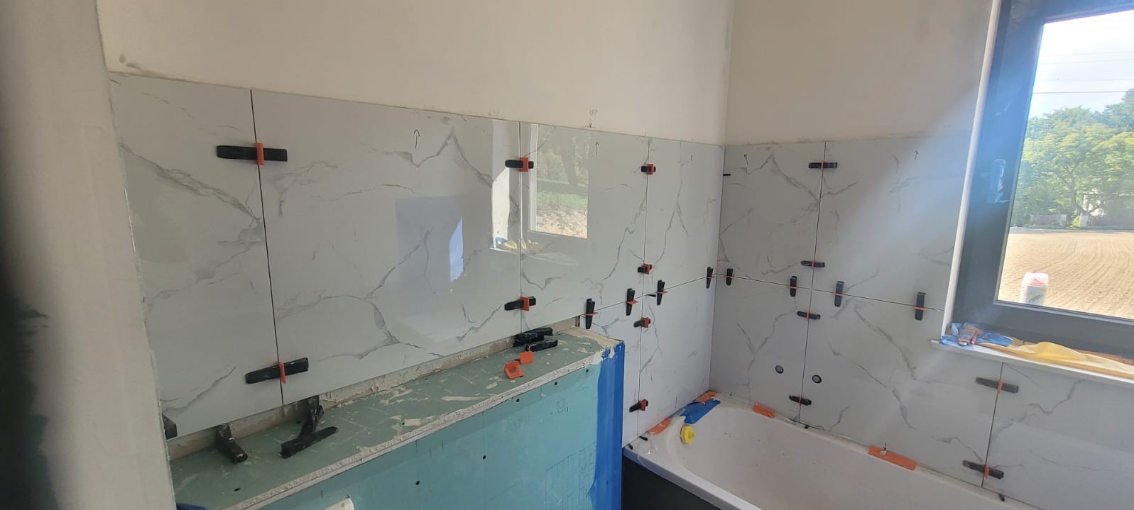 Świeżo ułożone, błyszczące płytki imitujące marmur na ścianach łazienki, z użyciem poziomujących klipsów i klinów, widoczna wanna i okno z widokiem na pole.