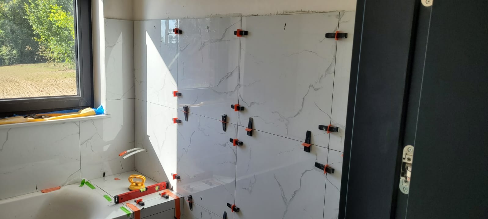 Świeżo ułożone, białe płytki imitujące marmur na ścianie łazienki, zabezpieczone pomarańczowymi i czarnymi klipsami poziomującymi, widoczne narzędzia glazurnicze, okno z widokiem na zewnątrz...