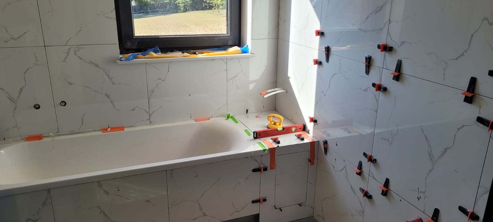 Łazienka w trakcie wykańczania: białe płytki imitujące marmur na ścianach, system poziomowania płytek, wanna obudowana płytkami, poziomica na wannie, okno w tle.