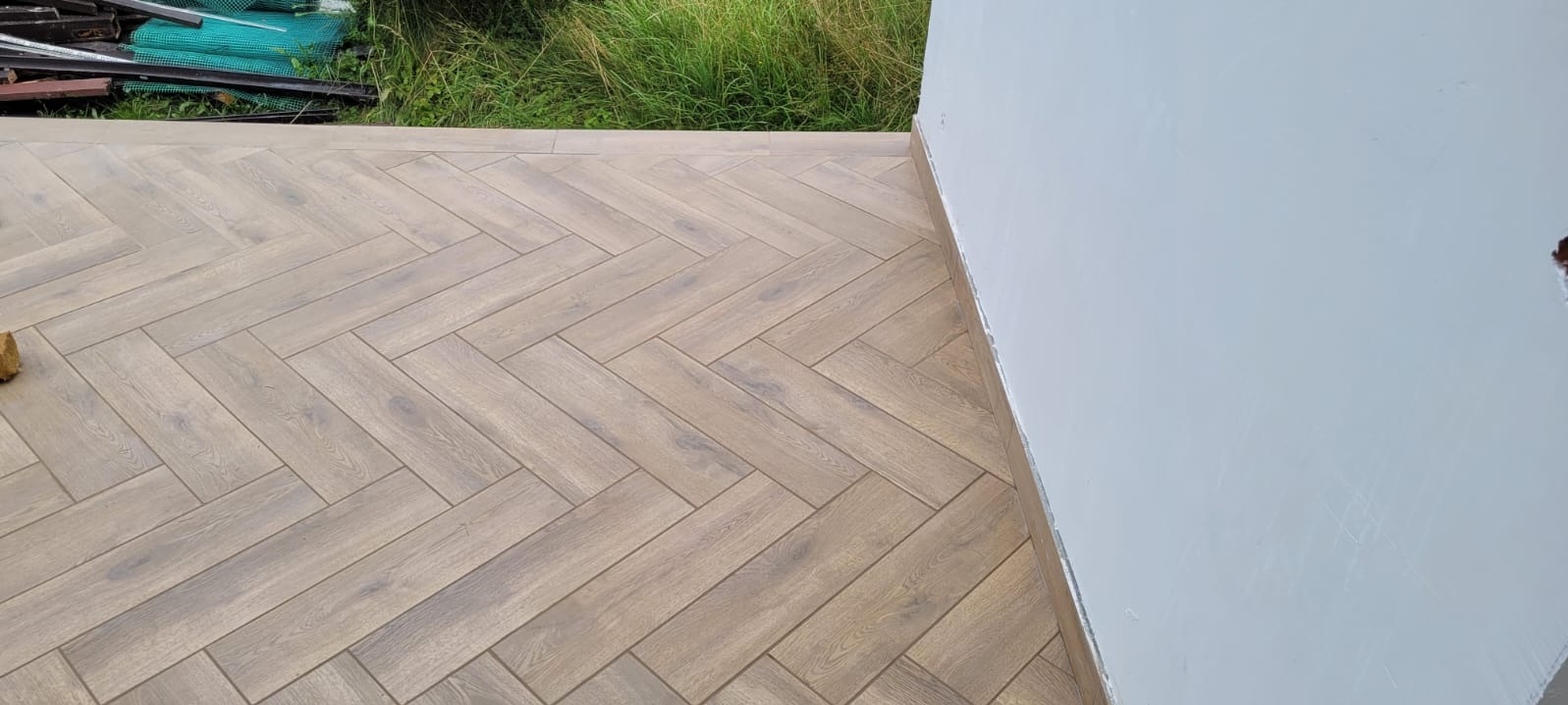 Ułożona pod kątem jodełki podłoga z paneli imitujących drewno, przy białej ścianie i zaroślach w tle.