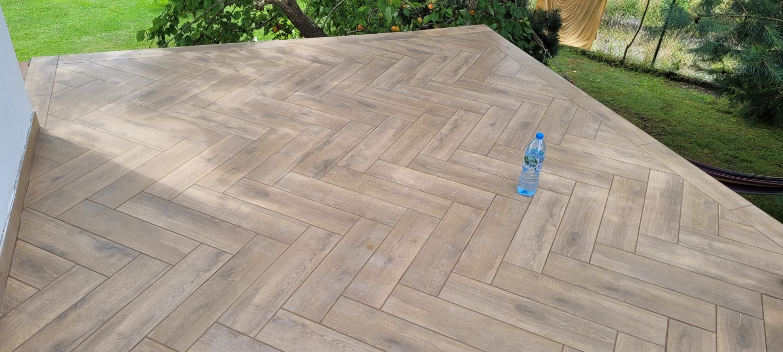Ułożona w jodełkę podłoga tarasu z płytek imitujących drewno, z butelką wody mineralnej w rogu, widok na trawnik i drzewa w tle.