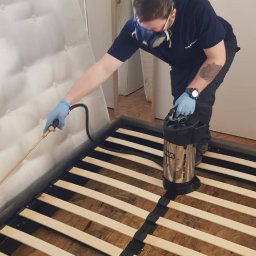 Pogotowie Domowe - Dezynsekcja ramy łóżka: pracownik w masce i rękawicach aplikuje preparat z metalowego opryskiwacza na drewnianą konstrukcję, obok materac.