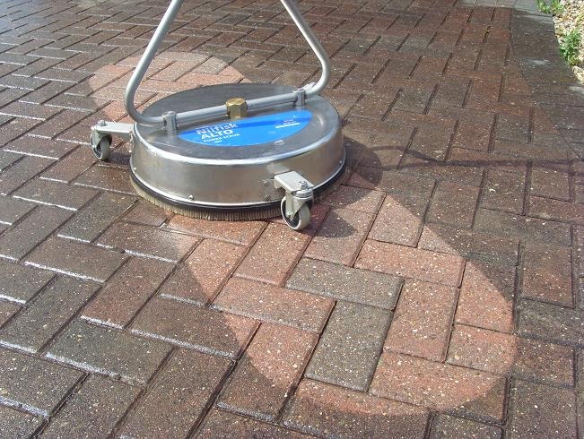 Rotacyjna szczotka czyszcząca Nilfisk Alto na kółkach, w trakcie pracy na kostce brukowej, widoczny efekt czyszczenia w postaci jaśniejszego okręgu na powierzchni.
