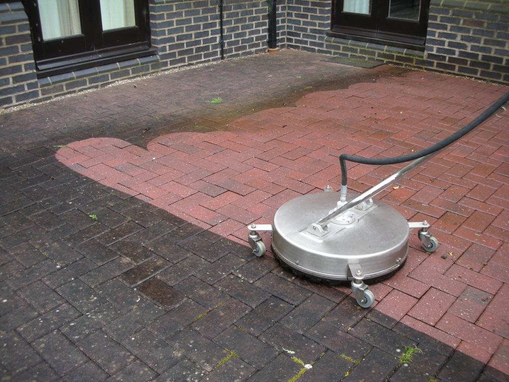 Czyszczenie kostki brukowej z użyciem myjki ciśnieniowej z okrągłą dyszą, widoczny kontrast między czystą, mokrą kostką a brudną, suchą powierzchnią, ceglany budynek w tle.