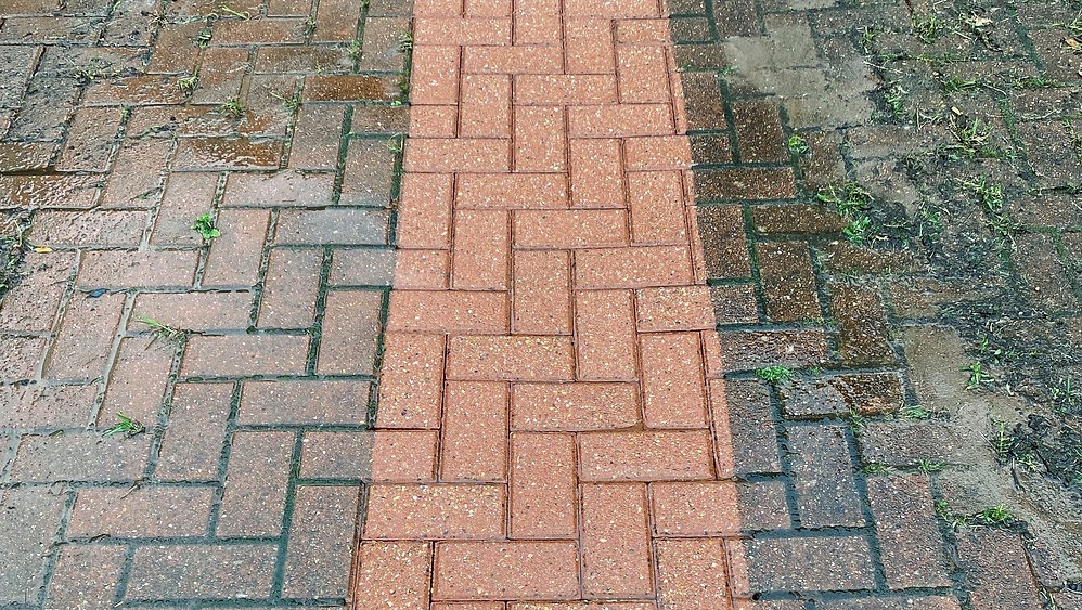 Częściowo umyta kostka brukowa ułożona w jodełkę, widoczna różnica w kolorze i czystości między umytą a brudną częścią, trawa rosnąca między kostkami.