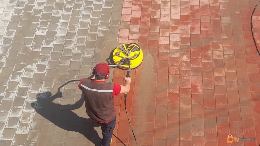 Pracownik w słuchawkach czyści czerwoną kostkę brukową za pomocą okrągłej myjki ciśnieniowej; widoczny kontrast między czystą i brudną powierzchnią.