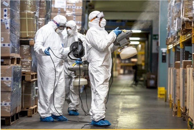 Pracownicy w kombinezonach ochronnych, maskach i rękawicach, używają zamgławiaczy elektrycznych w magazynie z regałami wypełnionymi towarami.