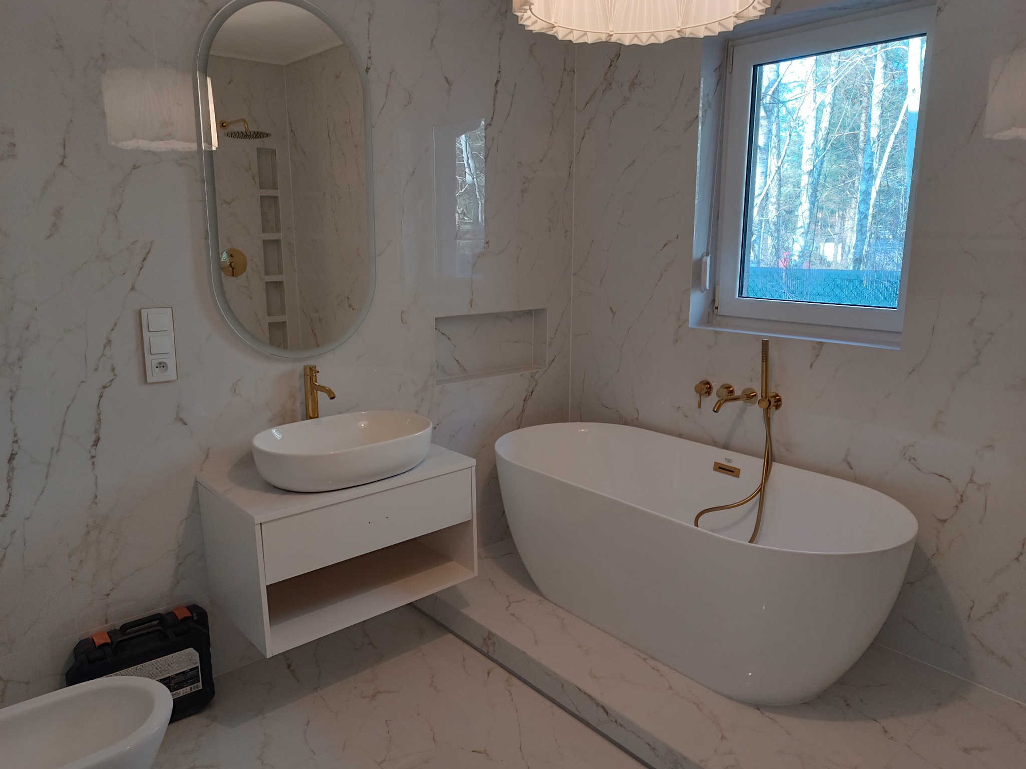 Nowoczesna łazienka z wanną wolnostojącą, umywalką nablatową i lustrem w owalnej ramie. Marmurowe płytki na ścianach i podłodze. Złota armatura. Widok z okna na drzewa.