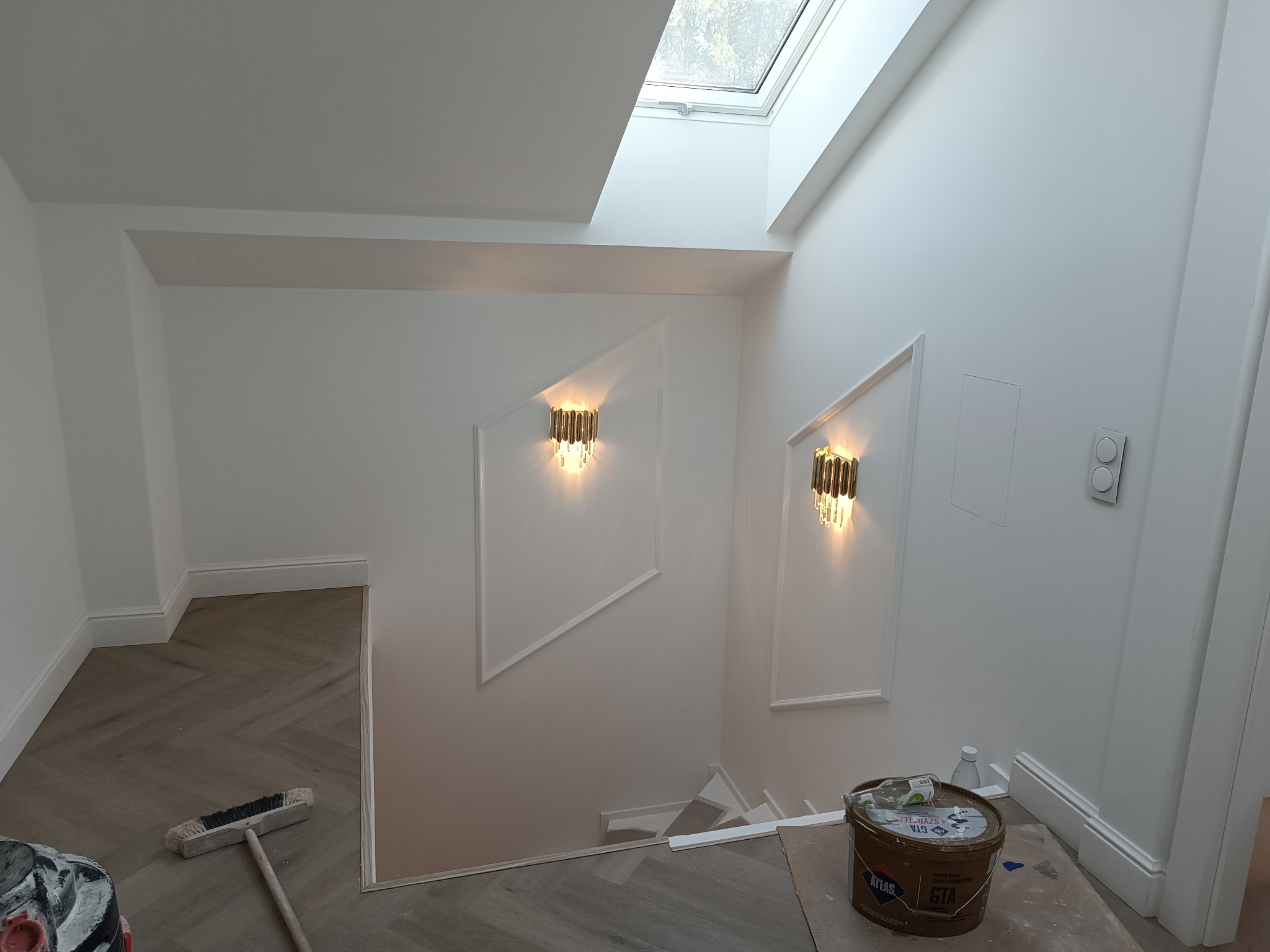 Białe wnętrze po malowaniu: ściany z dekoracyjnymi listwami, lampy ścienne, parkiet w jodełkę, widok na schody i okno dachowe. Wiadro z farbą i szczotka na podłodze.