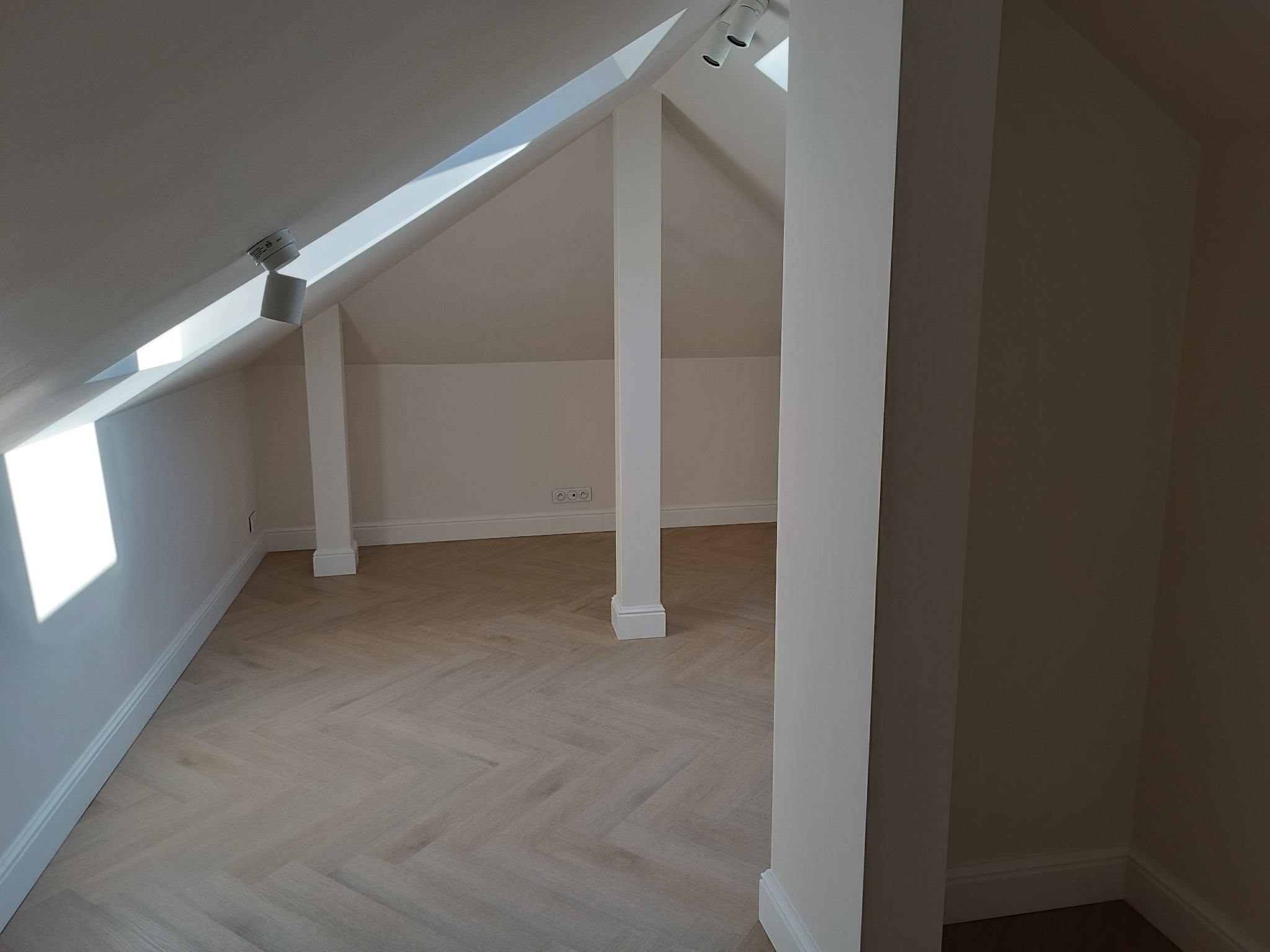 Jasne poddasze z parkietem w jodełkę, białe ściany i kolumny. Widoczne okno dachowe i oświetlenie punktowe. Gniazdka elektryczne na ścianie.
