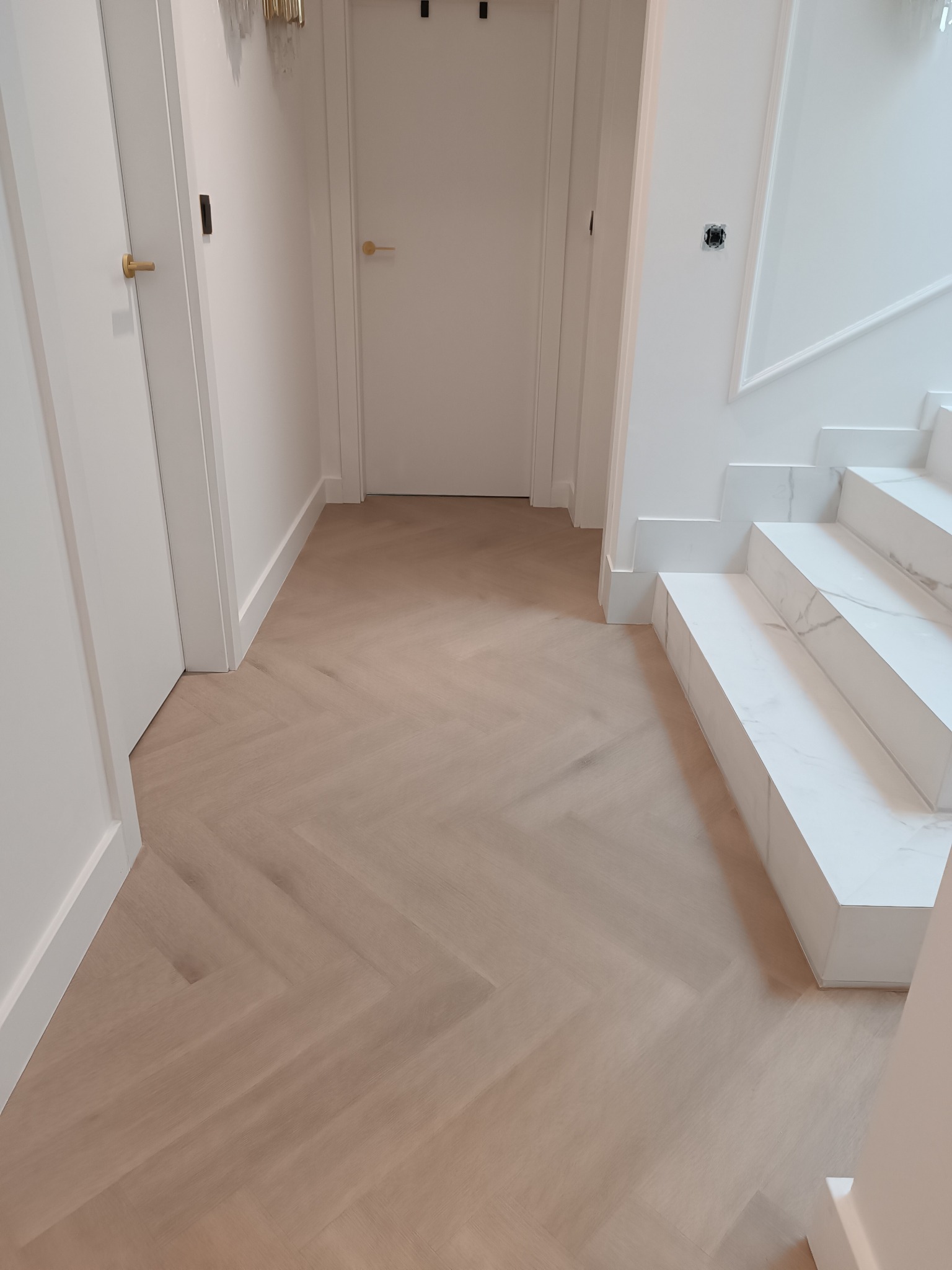 Jasne panele ułożone w jodełkę, białe drzwi z złotymi klamkami i marmurowe schody w nowoczesnym wnętrzu. Widok z góry, neutralne oświetlenie.