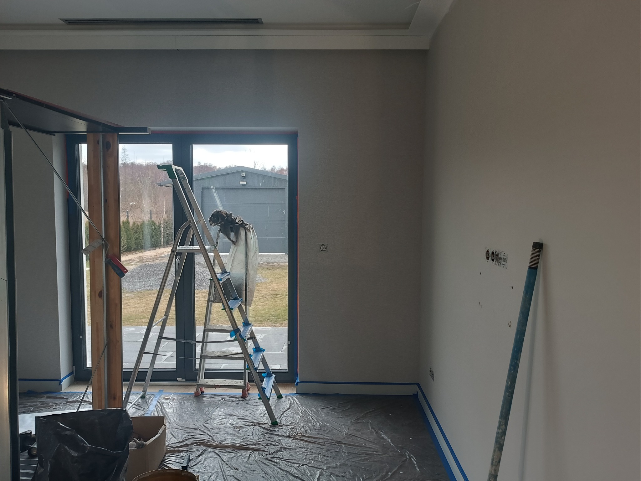 Przygotowania do malowania ścian w Markach: drabina, folia ochronna na podłodze, taśma malarska przy listwach. Świeżo pomalowana ściana w tle.