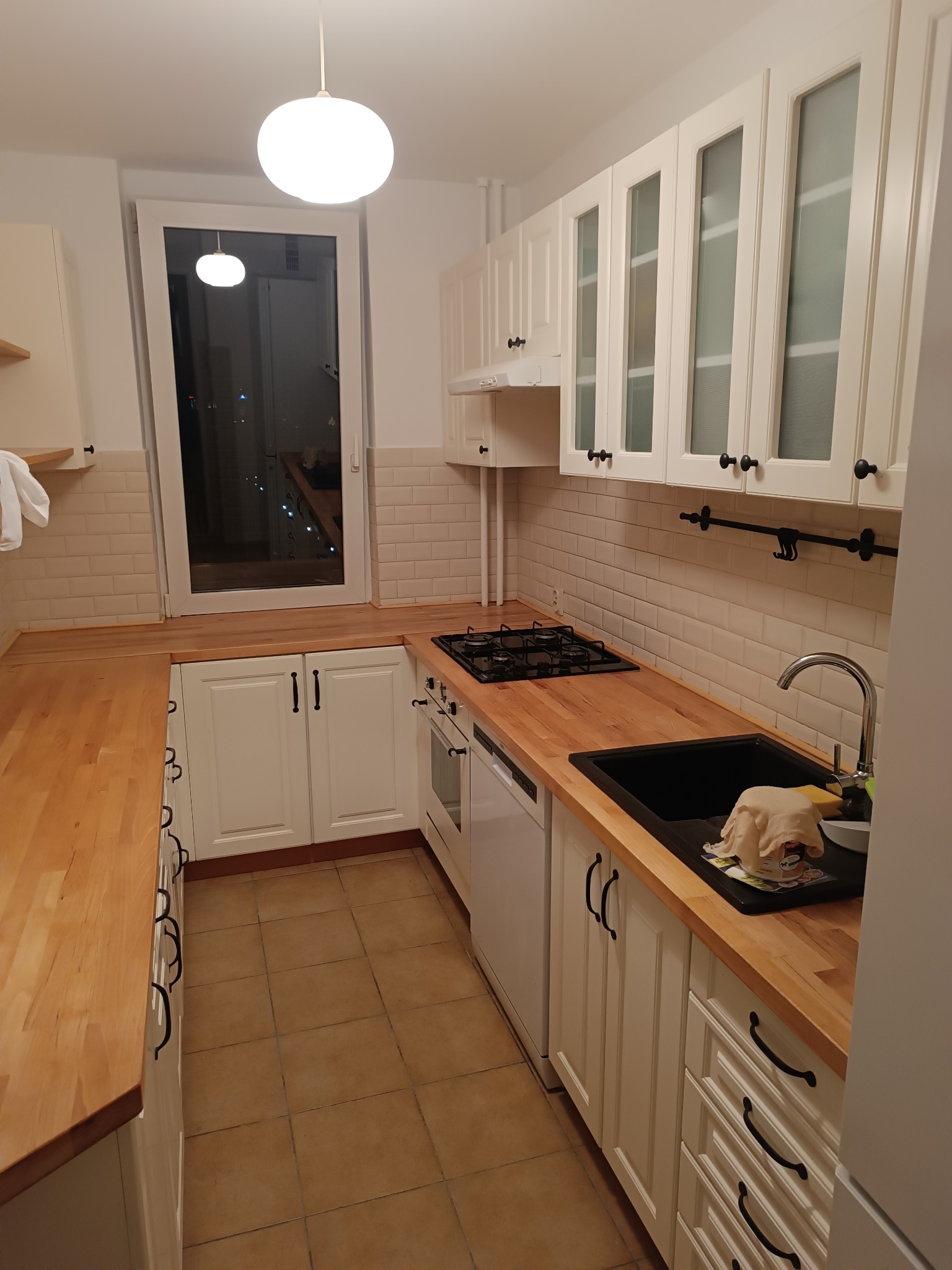 Jasna kuchnia w stylu skandynawskim z drewnianym blatem, białymi szafkami z czarnymi uchwytami i beżowymi płytkami na ścianie. Widoczny zlew, płyta gazowa i okno.