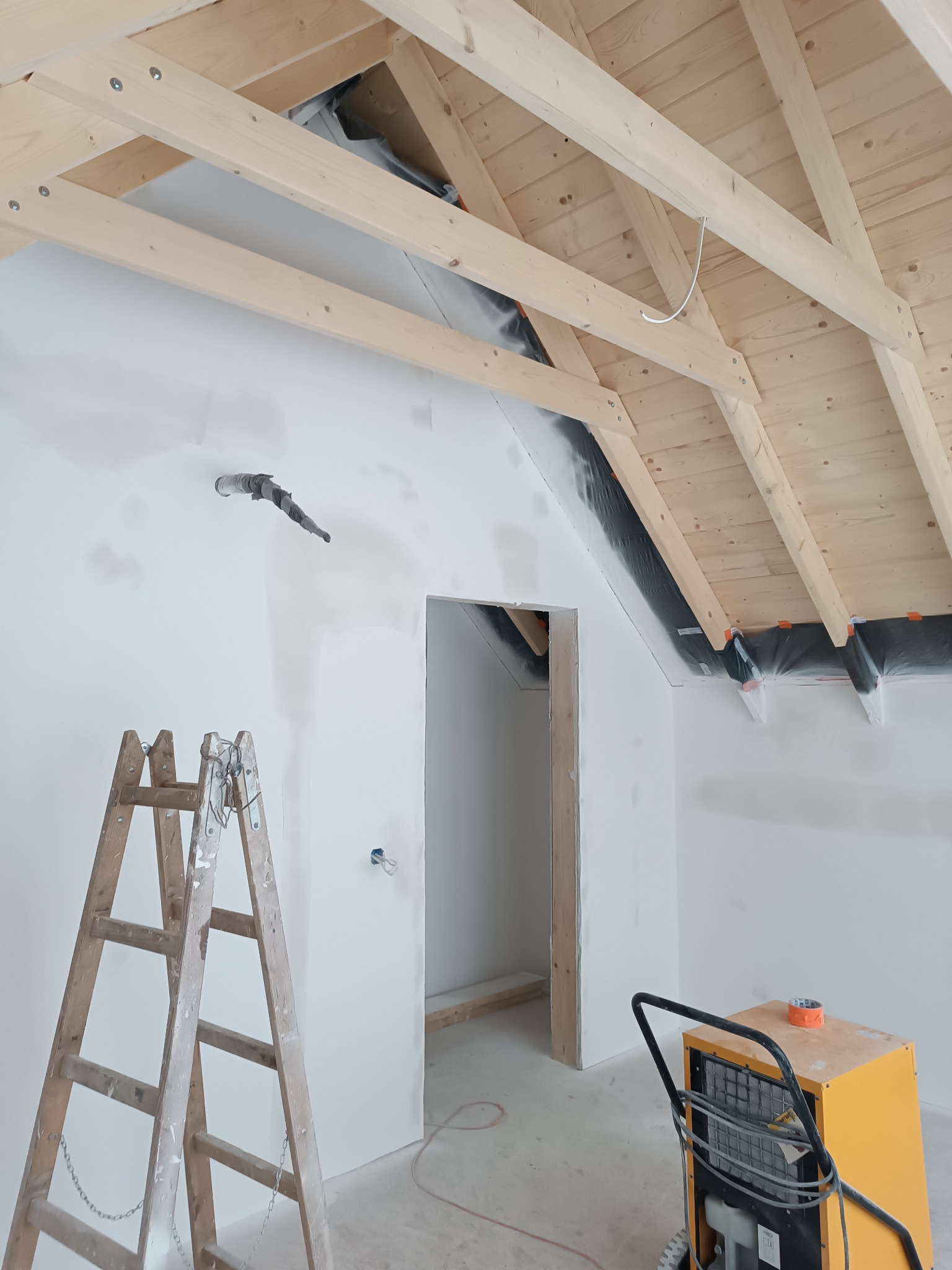 Poddasze w trakcie wykańczania: drewniana więźba dachowa, ściany po szpachlowaniu, drabina malarska i osuszacz powietrza. Widoczny otwór drzwiowy.