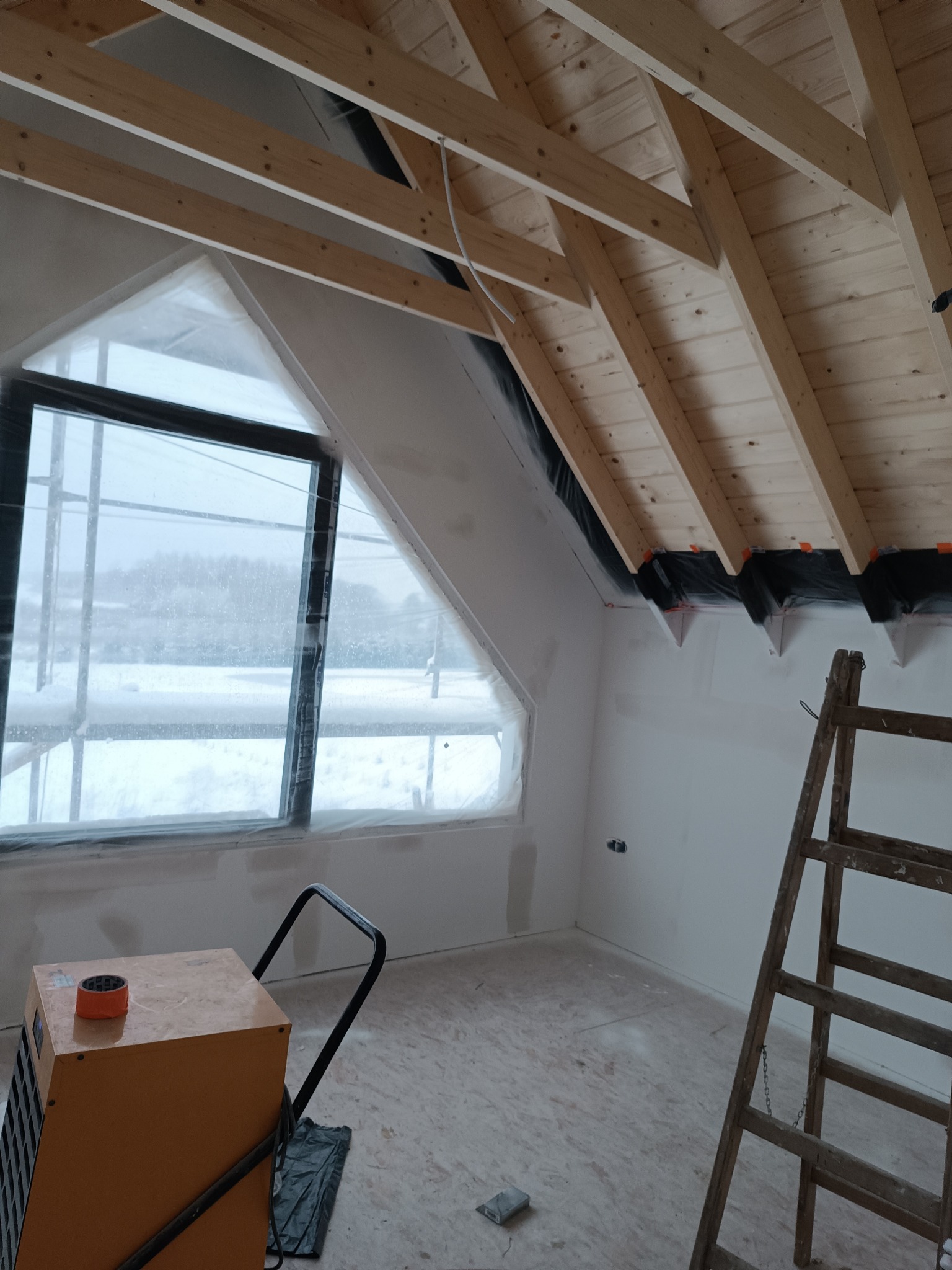 Poddasze w trakcie wykańczania: drewniana więźba dachowa, okno zabezpieczone folią, ściany po szpachlowaniu, drabina i osuszacz budowlany. Widok z dołu.