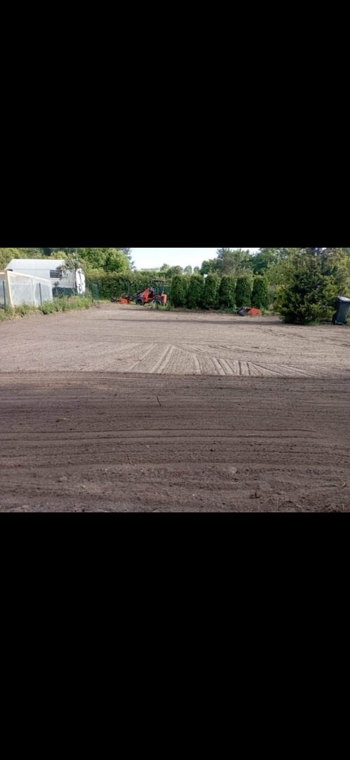 Przygotowany teren pod trawnik z widocznymi śladami po maszynach, w tle rząd tuj i maszyny rolnicze, po lewej stronie szklarnia.