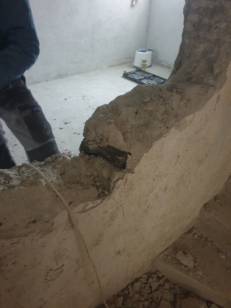Fragment ściany z widocznym otworem po wyburzeniu, z wystającymi elementami konstrukcyjnymi i instalacją elektryczną, w tle wiadro i skrzynka narzędziowa.