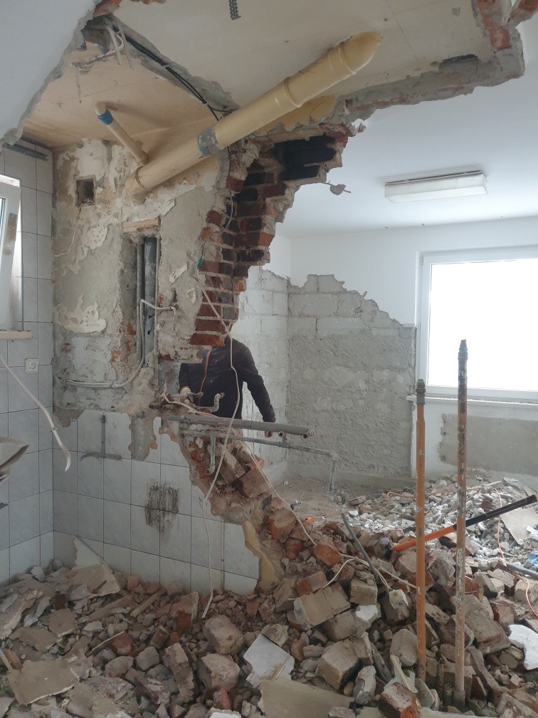 Wnętrze w trakcie demontażu ściany z widoczną instalacją hydrauliczną, fragmentami cegieł i gruzu na podłodze, zarys sylwetki pracownika w tle.