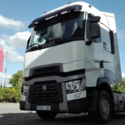 Biała ciężarówka Renault z widoczną tablicą rejestracyjną 5953 JBS, zaparkowana na placu, z logo Renault Trucks i flagami w tle, w słoneczny dzień.