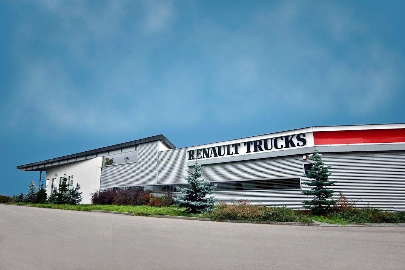 Budynek serwisu Renault Trucks z szarej blachy falistej, z napisem firmowym i czerwonym pasem na dachu, otoczony zielenią i młodymi drzewkami.