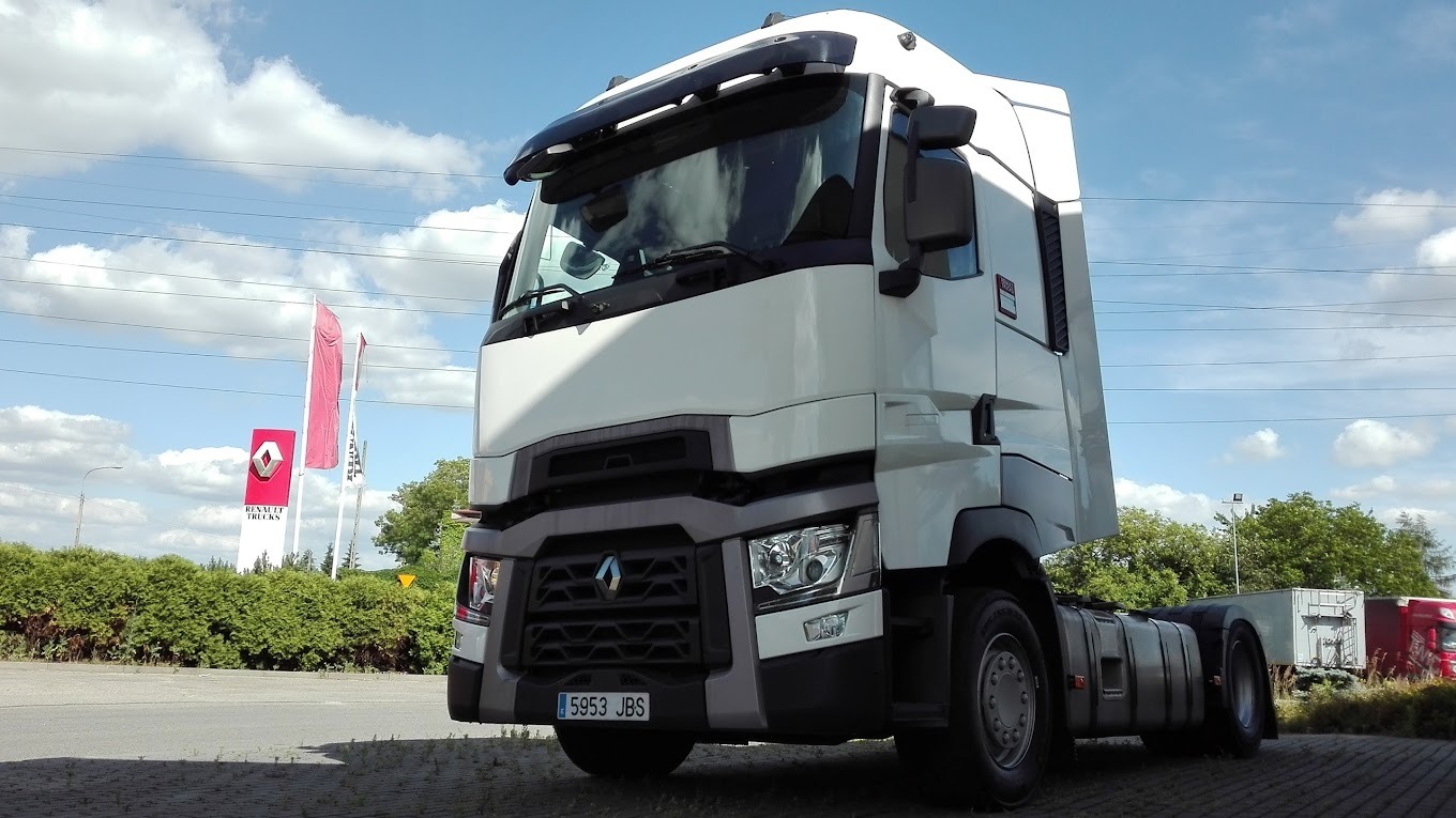 Biała ciężarówka Renault z widoczną tablicą rejestracyjną 5953 JBS, zaparkowana na placu, z logo Renault Trucks i flagami w tle, w słoneczny dzień.