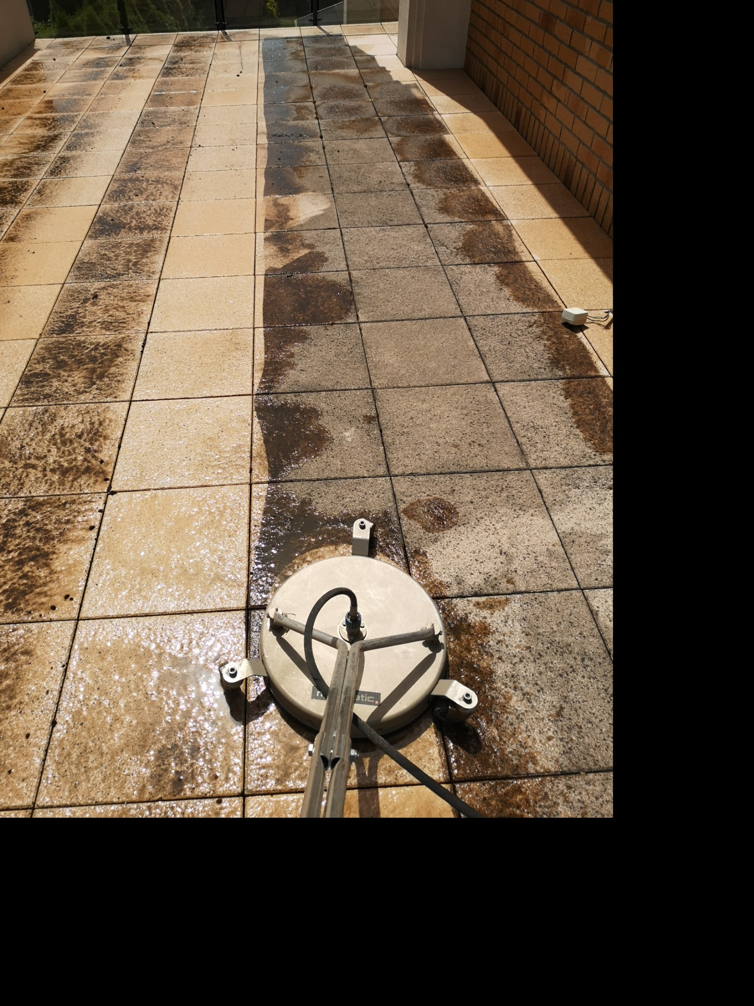 Czyszczenie kostki brukowej na tarasie za pomocą myjki ciśnieniowej z okrągłą dyszą, widoczna różnica między umytą a brudną powierzchnią, ceglana ściana w tle.