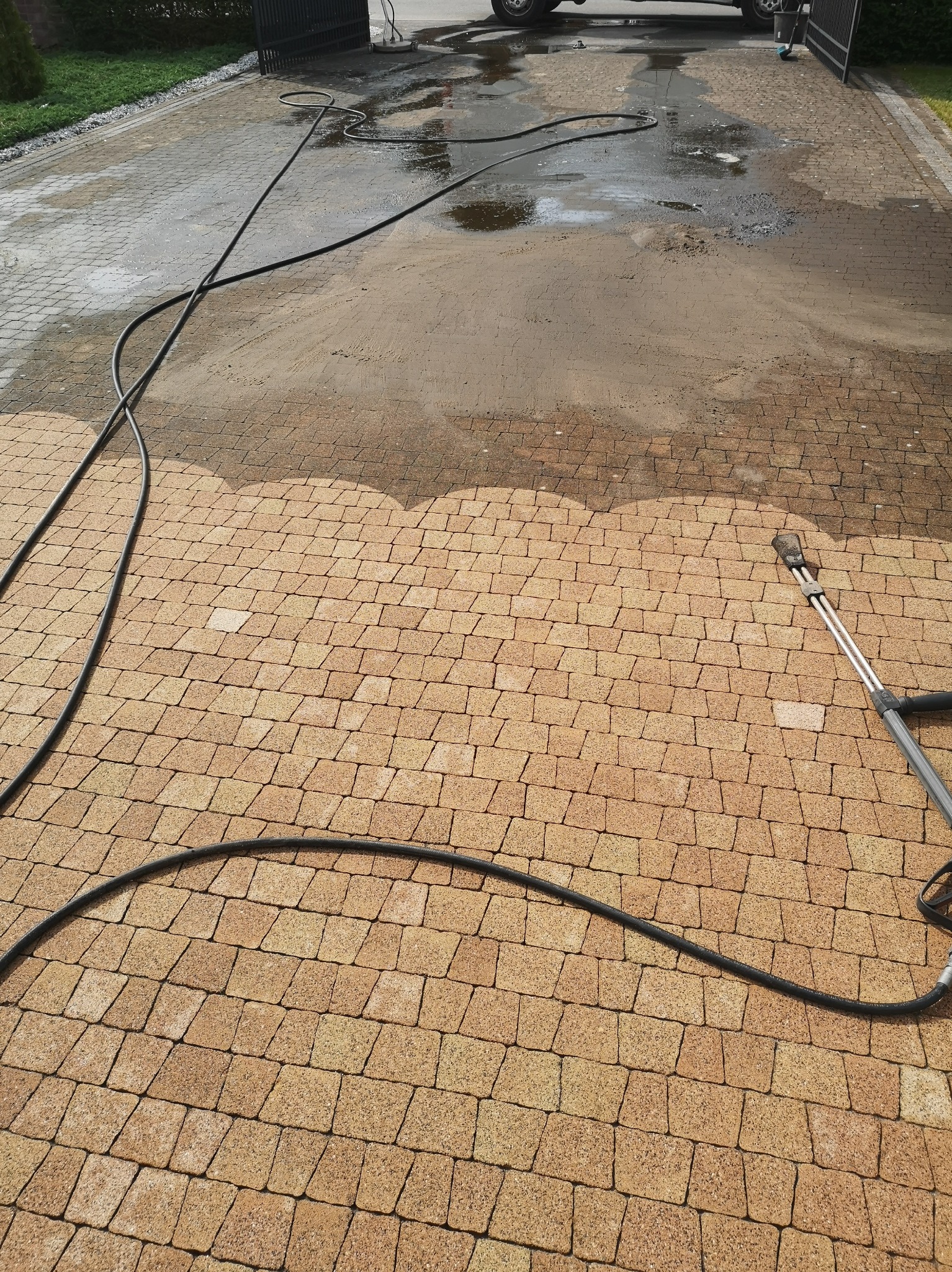 Częściowo wyczyszczona, brązowa kostka brukowa po myciu ciśnieniowym, z widocznym wężem i lancą myjki.