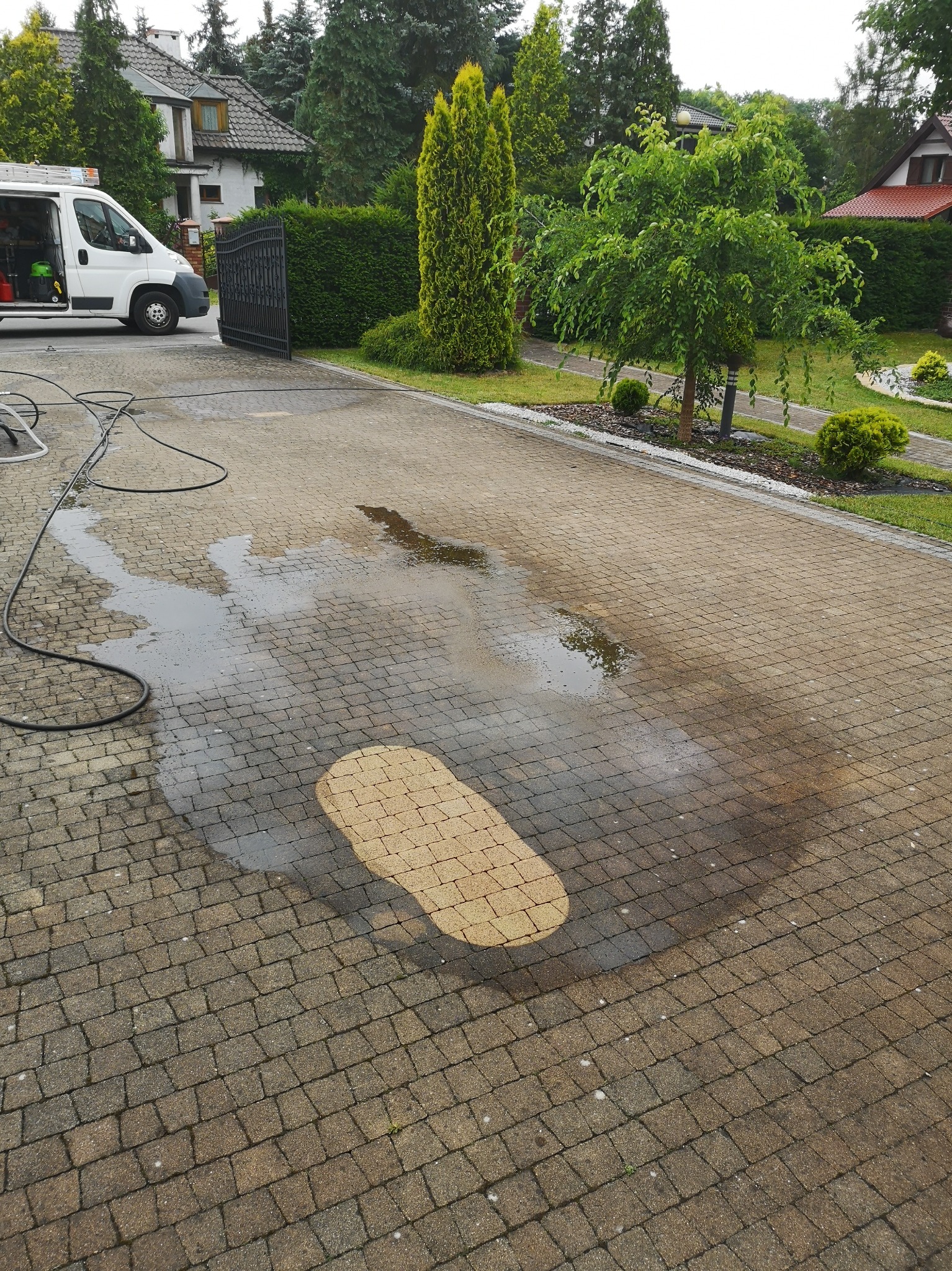 Mycie kostki brukowej na podjeździe domu jednorodzinnego, widoczny biały bus z wyposażeniem oraz ślady mokrej i czystej kostki.