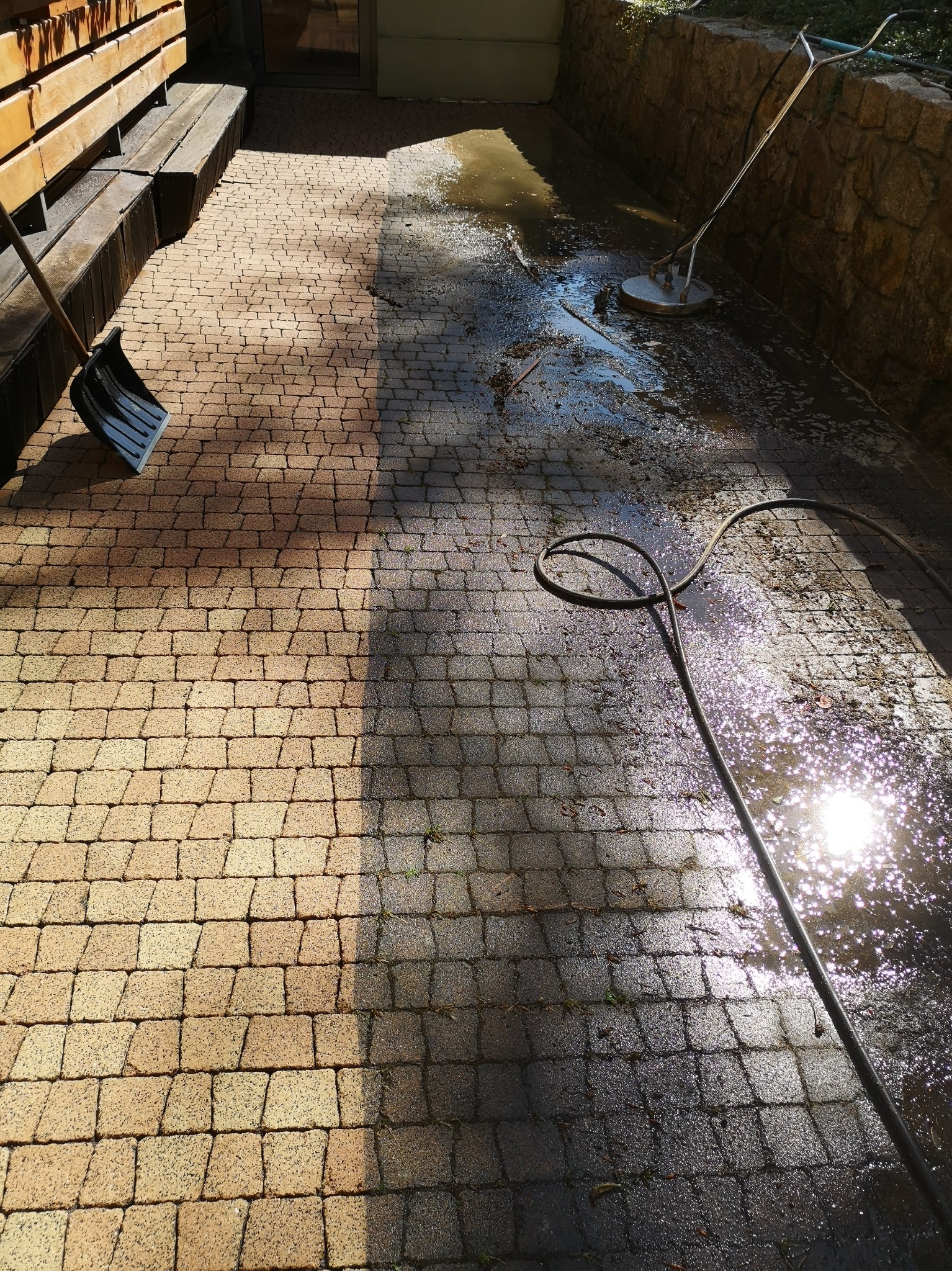 Częściowo wyczyszczona kostka brukowa z widoczną różnicą w kolorze, szpadel i myjka ciśnieniowa w tle.