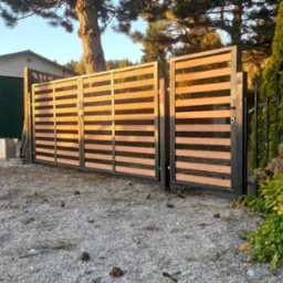 Nowoczesna brama wjazdowa z poziomymi panelami drewnopodobnymi w czarnej ramie, widoczna na tle żwirowego podjazdu i garażu z otwartymi drzwiami, z elementami roślinności przy ogrodzeniu.