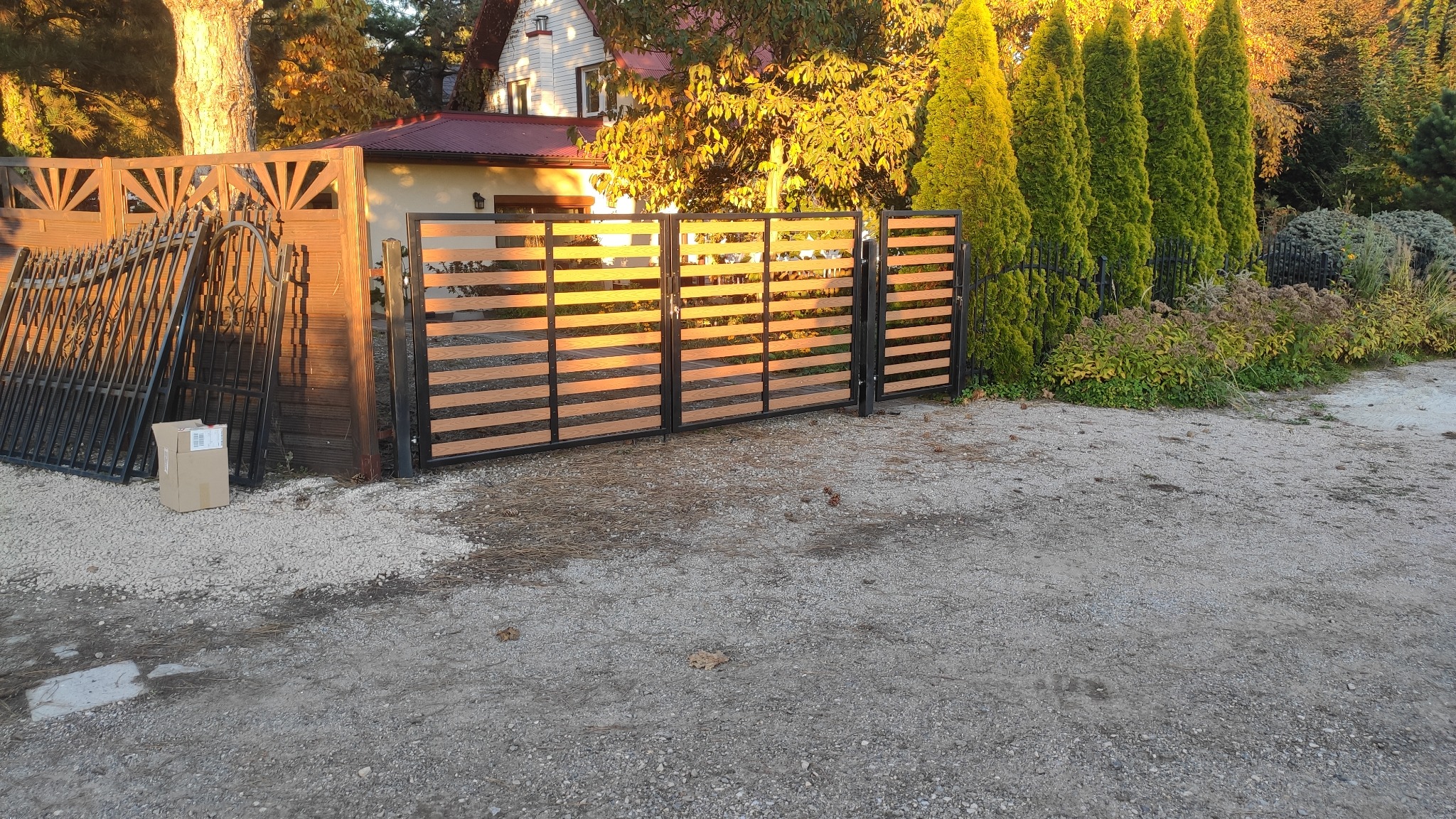 Nowoczesna brama wjazdowa z poziomymi, drewnopodobnymi panelami, widoczne elementy montażowe i nieukończone fragmenty ogrodzenia obok, dom w tle.