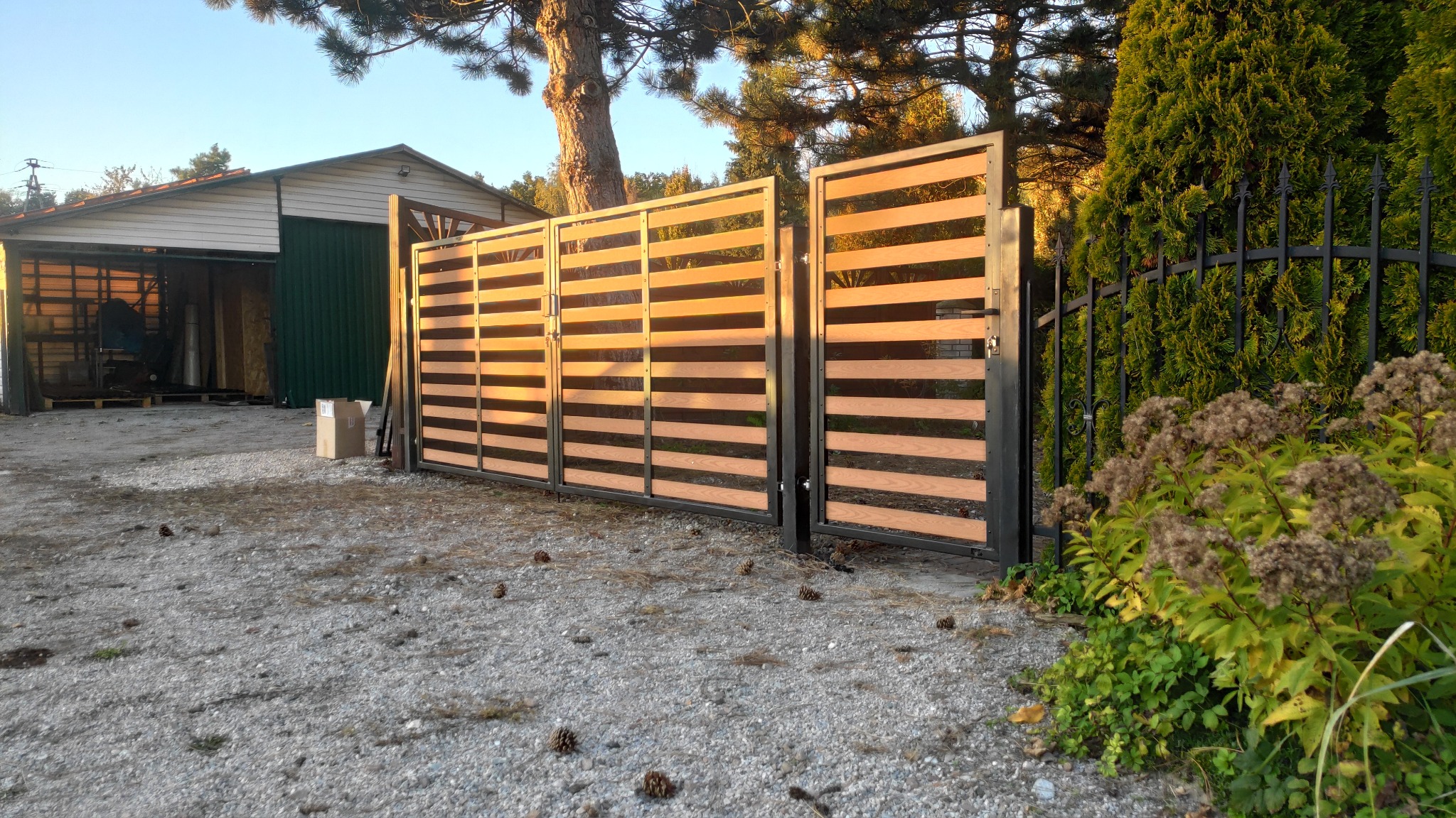 Nowoczesna brama wjazdowa z poziomymi panelami drewnopodobnymi w czarnej ramie, widoczna na tle żwirowego podjazdu i garażu z otwartymi drzwiami, z elementami roślinności przy ogrodzeniu.