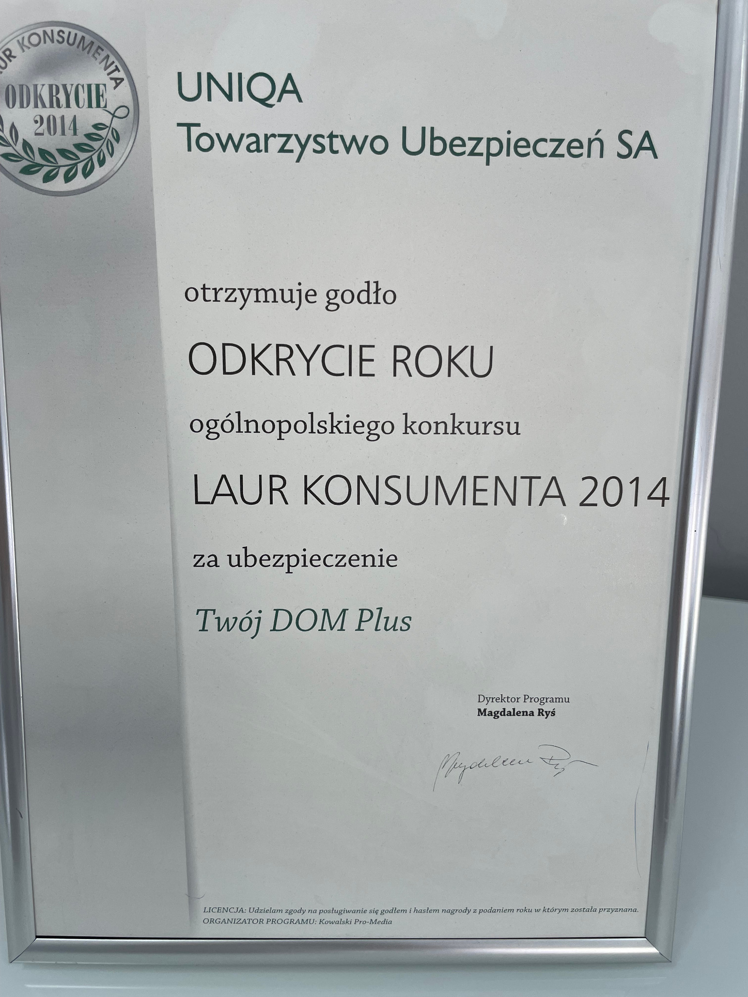 Certyfikat 'Odkrycie Roku Laur Konsumenta 2014' dla UNIQA Towarzystwo Ubezpieczeń SA za ubezpieczenie 'Twój DOM Plus', podpisany przez Dyrektor Programu Magdalenę Ryś, w srebrnej ramce.