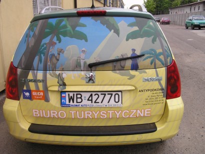 Żółty samochód Peugeot 307 SW z oklejoną tylną szybą przedstawiającą pustynny krajobraz z palmami, wielbłądami i postaciami, reklama biura turystycznego 'Biuro Turystyczne Antypodkowa' z numerem...